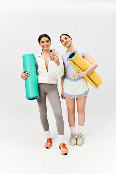 Two pretty teenage girls, one brunette, standing together in sportive attire holding yoga mats on grey studio background. — Stock Photo