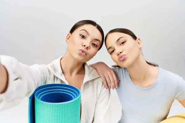Двоє красивих друзів-підлітків, одна брюнетка, позують з килимком для йоги, виключаючи спокій у спортивному вбранні на сірому тлі. — стокове фото