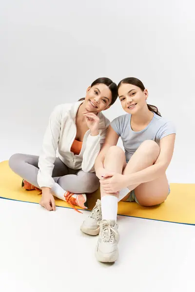 Deux jolies adolescentes brunes en tenue de sport assises sur un tapis, prenant une pose pour la caméra dans un studio. — Photo de stock
