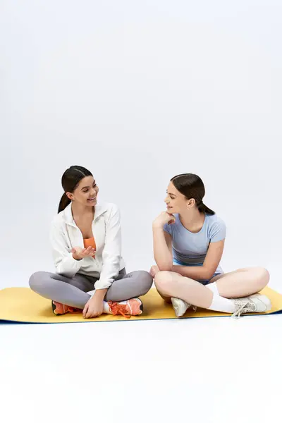 Две симпатичные брюнетки-девочки в спортивном костюме сидят на коврике для йоги, разговаривая и общаясь в спокойной студии. — стоковое фото