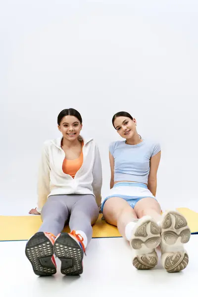 Two pretty and brunette teenage girls in sportive attire sit together on a mat with their feet up in a studio setting. — Stock Photo