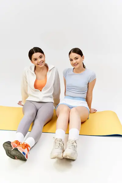 Две симпатичные брюнетки-девочки в спортивном наряде сидят на коврике со скрещенными ногами в студии. — стоковое фото