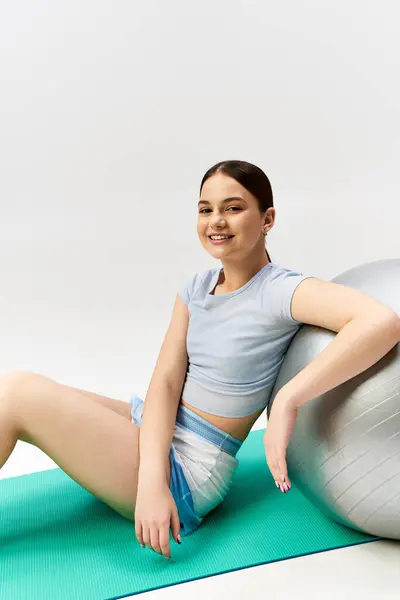 A pretty, brunette teenage girl in sporty attire gracefully balances on a yoga ball in a studio — Stock Photo