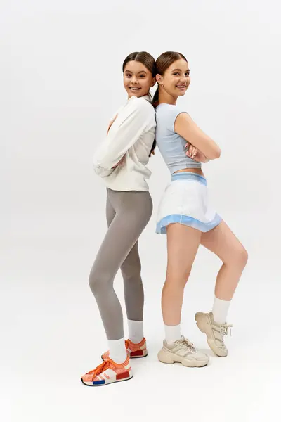 Two pretty, athletic teenage girls, brunette and in sportive attire, stand side by side against a grey studio background. — Stock Photo