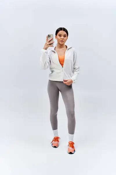 Une fille brune en tenue de sport prend joyeusement un selfie avec son téléphone portable dans un cadre studio. — Photo de stock