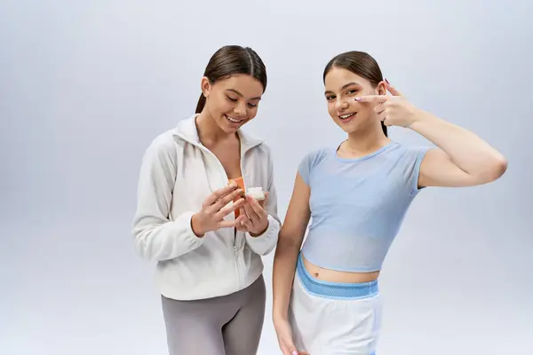 Duas adolescentes bonitas e morenas em trajes esportivos ficam ao lado uma da outra com uma pose confiante em um fundo de estúdio cinza. — Fotografia de Stock