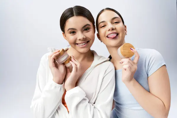Two pretty and brunette teenage girls in sportive attire strike a pose while holding cream in a studio on a grey background. — Stock Photo
