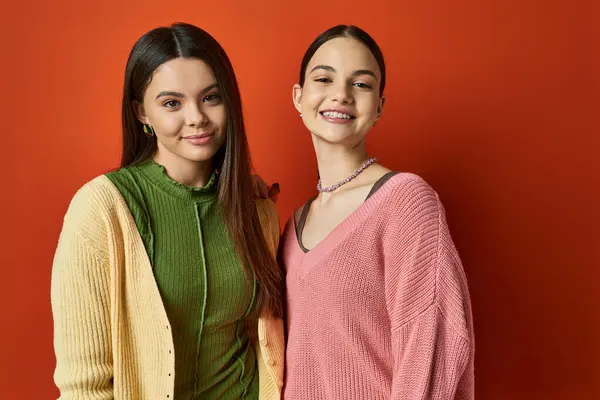 Two pretty and brunette teenage girls in casual attire standing next to each other in front of a vibrant red wall. — Stock Photo