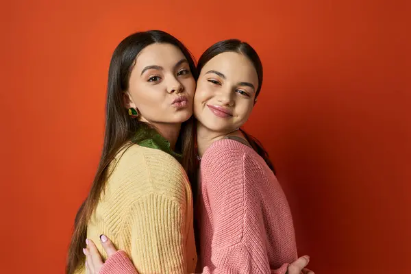 Дві красиві брюнетки дівчата-підлітки в повсякденному вбранні ніжно обіймають один одного перед яскравою червоною стіною. — стокове фото