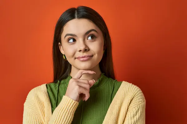 A brunette woman in casual attire looks thoughtful with her hand on her chin, standing against an orange background. — Stock Photo