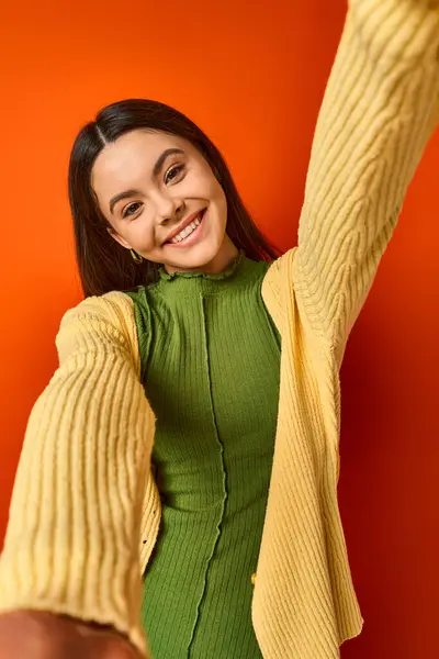 A brunette teenage girl wearing a green dress and a yellow cardigan poses elegantly on an orange background in a studio. — Stock Photo