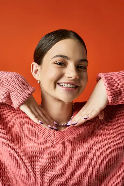 Uma adolescente morena em um suéter rosa está sorrindo brilhantemente contra um fundo laranja em um estúdio. — Fotografia de Stock