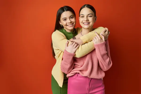 Two pretty, brunette teenage girls in casual attire embrace each other affectionately in a heartfelt moment in front of a red background. — Stock Photo