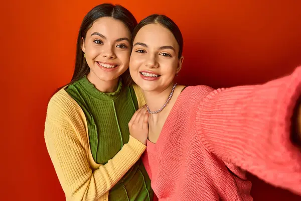 Two young women, brunette and pretty, hug each other in front of a red wall in a heartwarming display of friendship. — Stock Photo