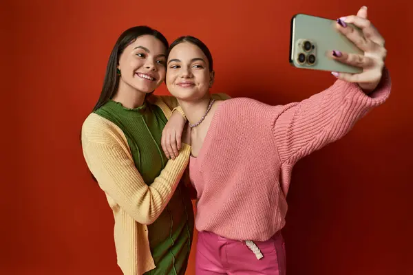 Dos bonitos amigos adolescentes capturando un momento con un teléfono celular en un estudio sobre un fondo naranja. - foto de stock