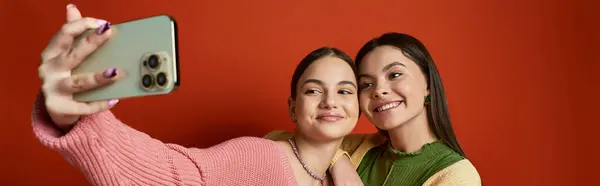 Two young women, friends, taking a selfie with a smartphone against an orange background in a studio setting. — Stock Photo