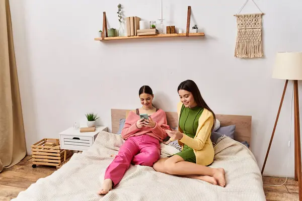 Дві молоді жінки в повсякденному одязі, сидячи на ліжку, занурені в мобільний телефон. — стокове фото