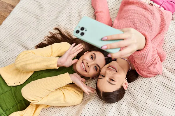 Deux jeunes femmes habillées de façon désinvolte, partageant un moment de rire alors qu'elles étaient allongées sur un lit, absorbées dans un téléphone portable. — Photo de stock