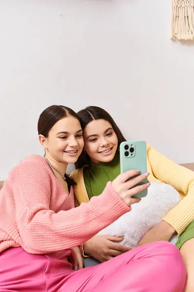 Two pretty teenage girls in casual attire sitting on a couch, capturing a fun moment by taking a selfie together. — Stock Photo