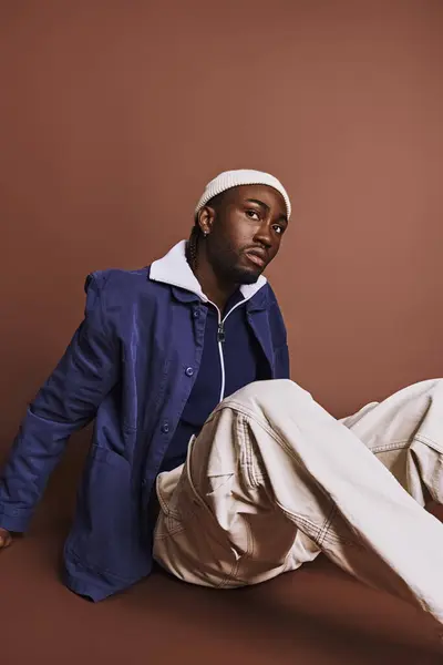Стильний афроамериканський чоловік сидить на землі в синій куртці і білих штанях. — стокове фото