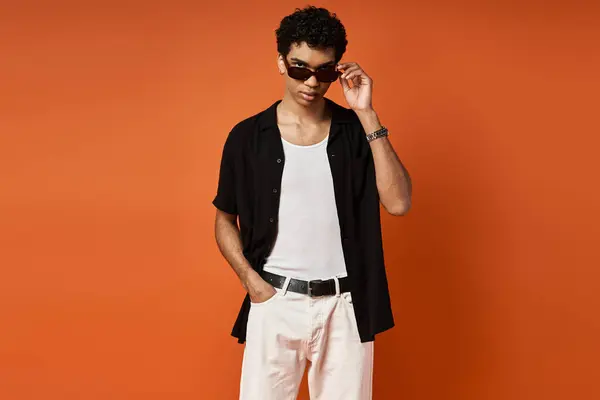 Homem bonito com óculos de sol em camisa preta e calças brancas marcando uma pose contra o fundo laranja vibrante. — Fotografia de Stock