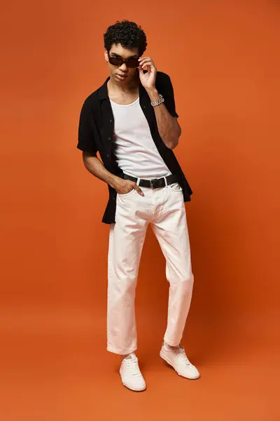 Bonito homem de calças brancas e camisa preta posa em vibrante pano de fundo laranja. — Fotografia de Stock