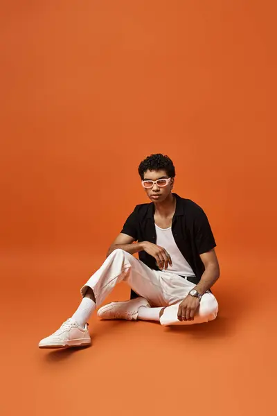 Handsome African American man sitting down wearing stylish sunglasses on a bright orange background. — Stock Photo