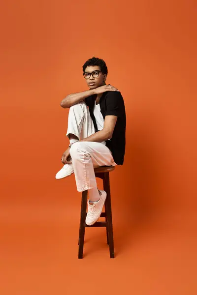 A handsome African American man sits on a stool in front of a bright orange background. — Stock Photo