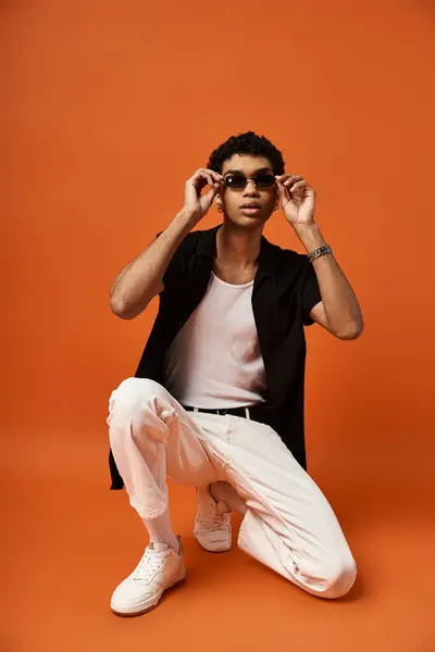 Handsome man in white pants and sunglasses crouching on orange background. — Stock Photo