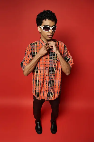 Hombre afroamericano guapo con camisa naranja y gafas de sol posando sobre fondo rojo. - foto de stock