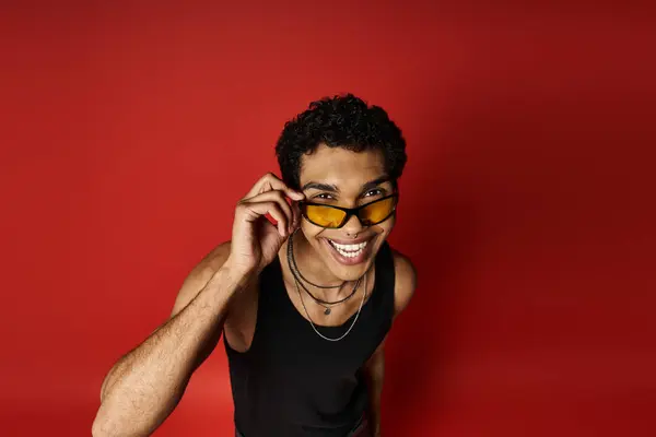 Un hombre afroamericano guapo lleva con confianza gafas de sol sobre un fondo rojo audaz. — Stock Photo