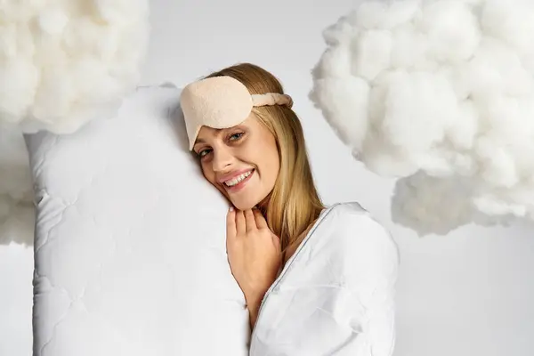 Eine verträumte blonde Frau im kuscheligen Pyjama hält ein Kissen, umgeben von flauschigen Wolken. — Stockfoto