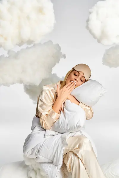 Una mujer rubia soñadora en pijama acogedor se sienta pacíficamente sobre nubes esponjosas. - foto de stock