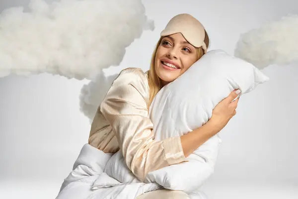 Femme blonde rêveuse en pyjama confortable assis au milieu des nuages avec un oreiller. — Photo de stock