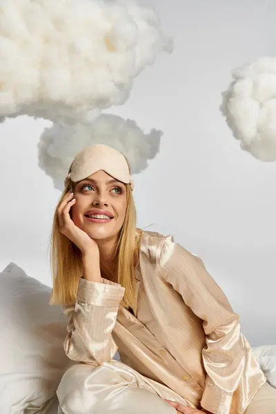 Eine verträumte blonde Frau im kuscheligen Pyjama sitzt auf einem weißen Kissen zwischen flauschigen Wolken. — Stockfoto