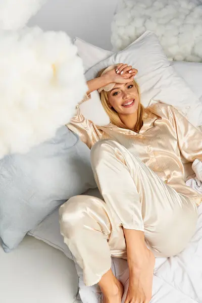 Une femme blonde rêveuse en pyjama confortable repose sur un lit à côté d'oreillers. — Photo de stock