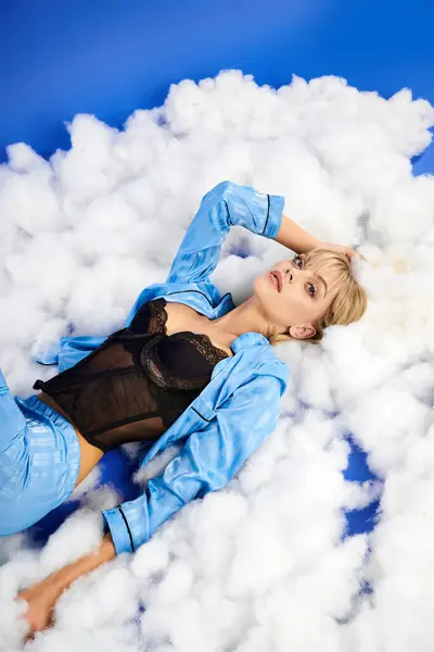 Una mujer rubia cautivadora vestida con un atuendo vibrante yace sobre un montón de nubes esponjosas contra un cielo azul. - foto de stock