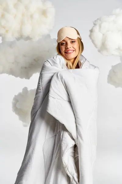 Femme blonde en pyjama confortable, drapée d'une couverture blanche, posant dans des nuages pelucheux. — Photo de stock