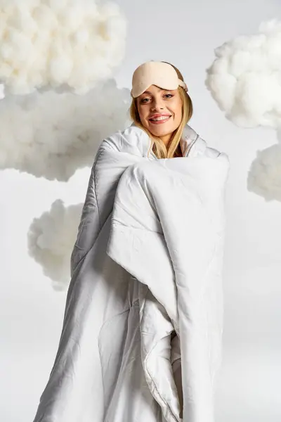 Blonde woman in cozy pyjamas, draped in white blanket, posing in fluffy clouds. — Stock Photo