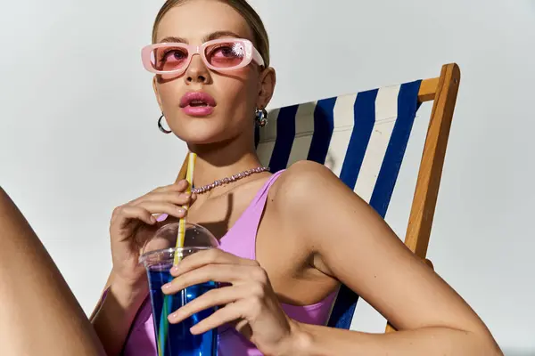 Mulher elegante usando óculos de sol, segurando uma bebida na cadeira de praia. — Fotografia de Stock