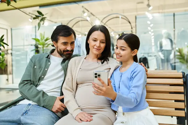 Une famille heureuse de quatre personnes est assise sur un banc, engloutie dans un écran de téléphone portable ensemble. — Photo de stock