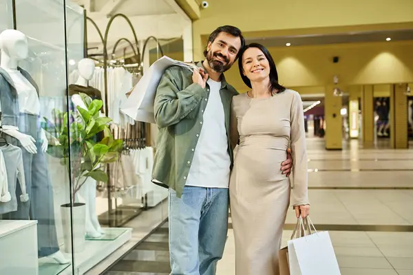 Pregnant couple excitedly holds shopping bags while exploring a bustling mall on a weekend outing. — Stock Photo