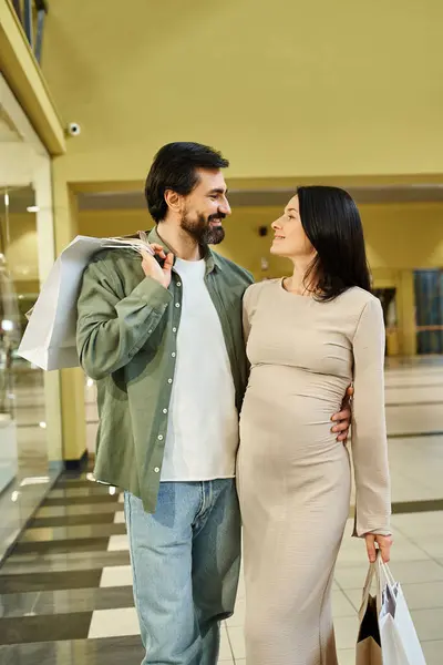 Una alegre pareja embarazada paseando por un centro comercial, sosteniendo bolsas llenas de golosinas. - foto de stock