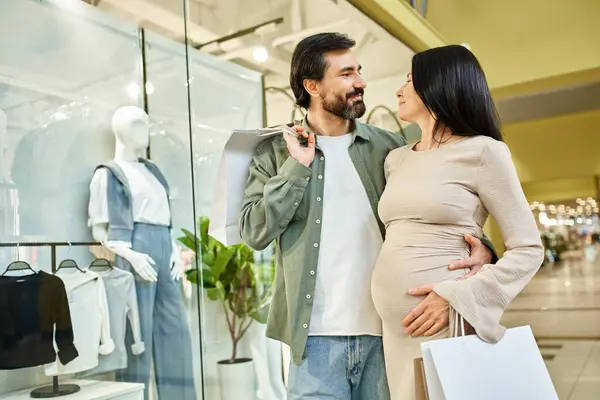 A pregnant couple joyfully holding shopping bags while strolling in a bustling shopping mall. — Stock Photo