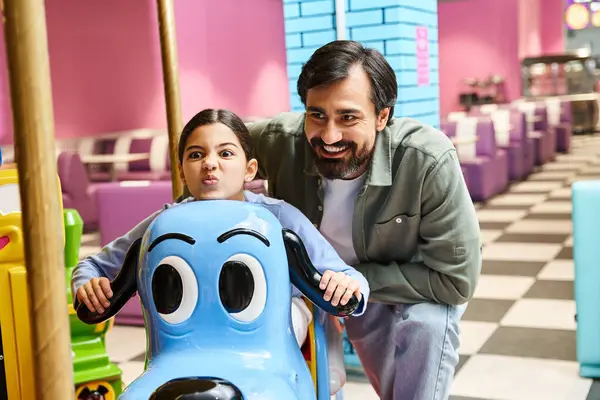 Père et enfant montent joyeusement un carrousel coloré dans un magasin de jouets, entouré d'autres familles heureuses dans une zone de jeu de centre commercial. — Photo de stock