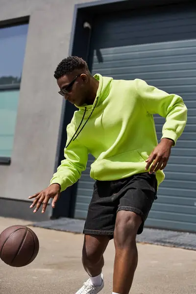 Hombre afroamericano guapo usa sudadera con capucha de neón y pantalones cortos, sosteniendo el baloncesto. - foto de stock