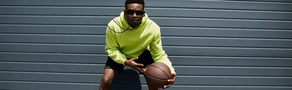 Guapo joven afroamericano con capucha verde sosteniendo una pelota de baloncesto. - foto de stock