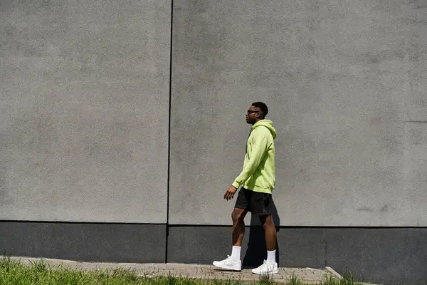 Stilvoller afroamerikanischer Mann in neongrünem Kapuzenpullover läuft an einer Wand vorbei. — Stockfoto