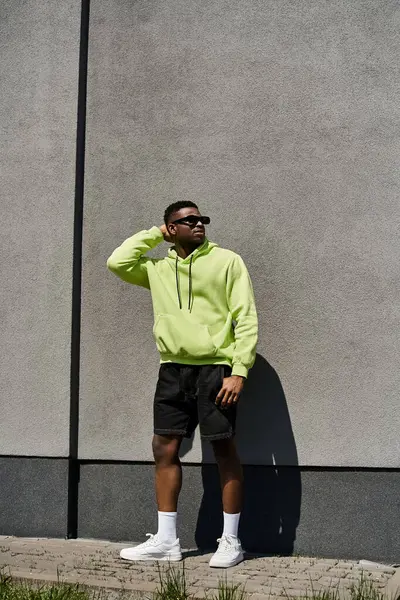 Stylischer Mann in neonfarbenem Kapuzenpulli und kurzer Hose lehnt an einer Wand. — Stockfoto