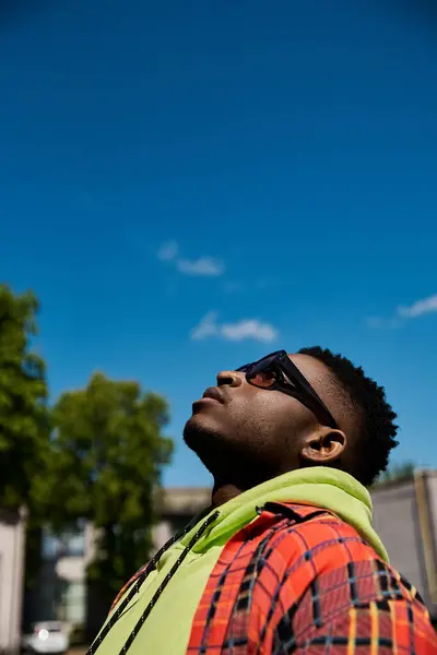 Junger Afroamerikaner in kariertem Kapuzenpulli und Sonnenbrille blickt in den Himmel. — Stockfoto